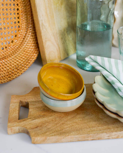 Stack of ceramic dipping bowls on a wooden cutting board with a glass of water and woven rattan charger plate in the background.