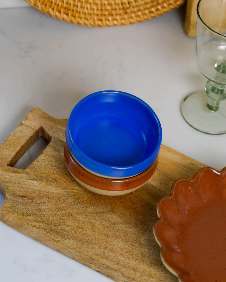 Blue stoneware dipping bowl on a wooden cutting board with a glass and woven rattan plate in the background