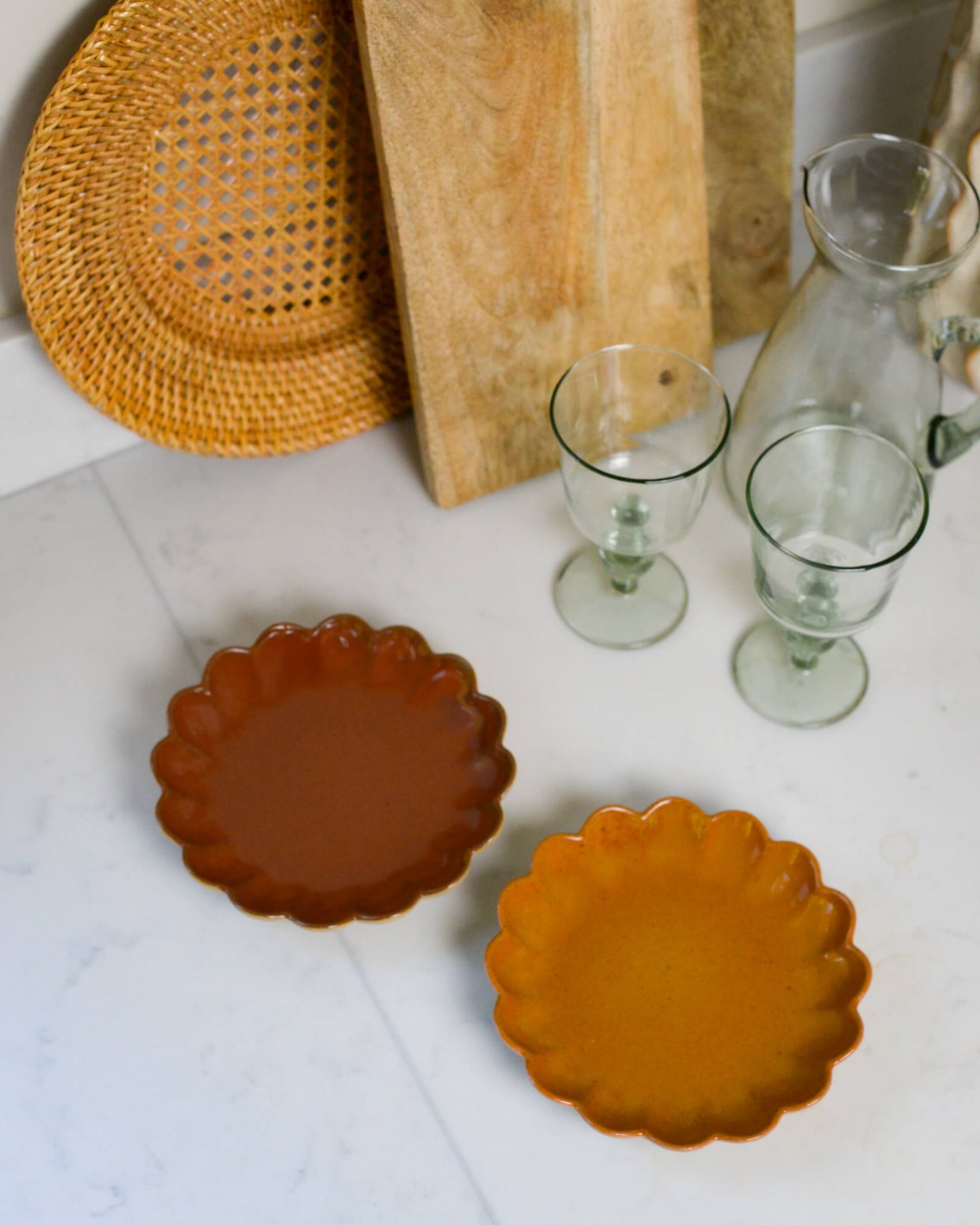 Two ceramic appetiser plates with scalloped edges on a white marble surface with a wooden cutting board and glassware in the background.
