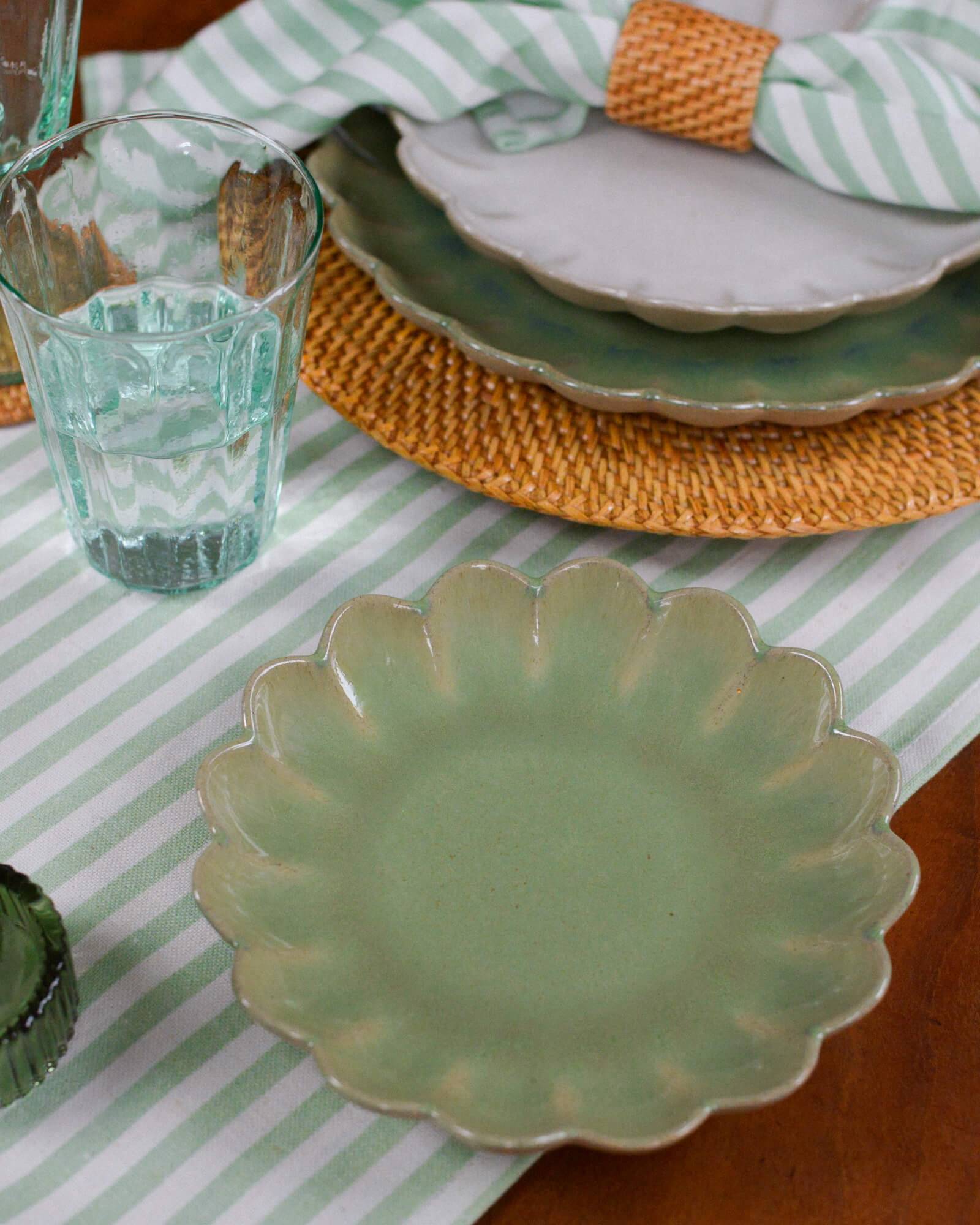 Green ceramic scalloped edged plate on a striped tablecloth with a glass of water and a woven rattan charger plate