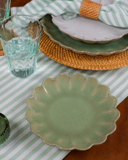 Green ceramic scalloped edged plate on a striped tablecloth with a glass of water and a woven rattan charger plate