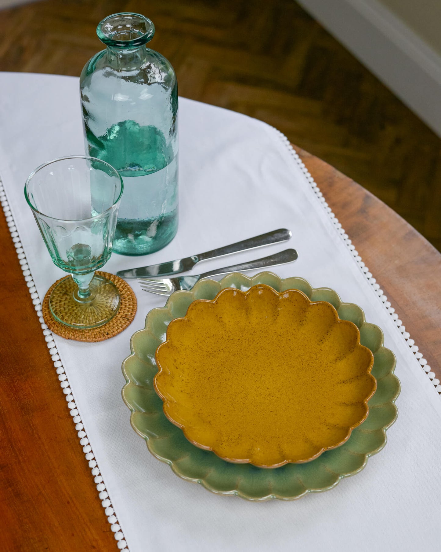 Dining table setting with a yellow scalloped edge plate, glass, and bottle on a white table runner.