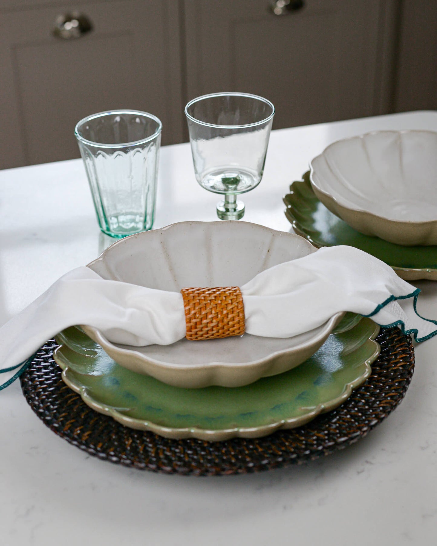 Dining table setting with stoneware scalloped edge plates, pasta bowls, glasses, and a woven placemat.