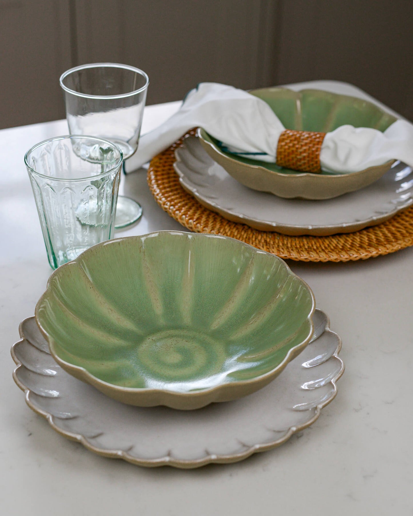 Dining table setting with green ceramic pasta bowl, white scalloped edge plates, and clear glasses on a neutral background.