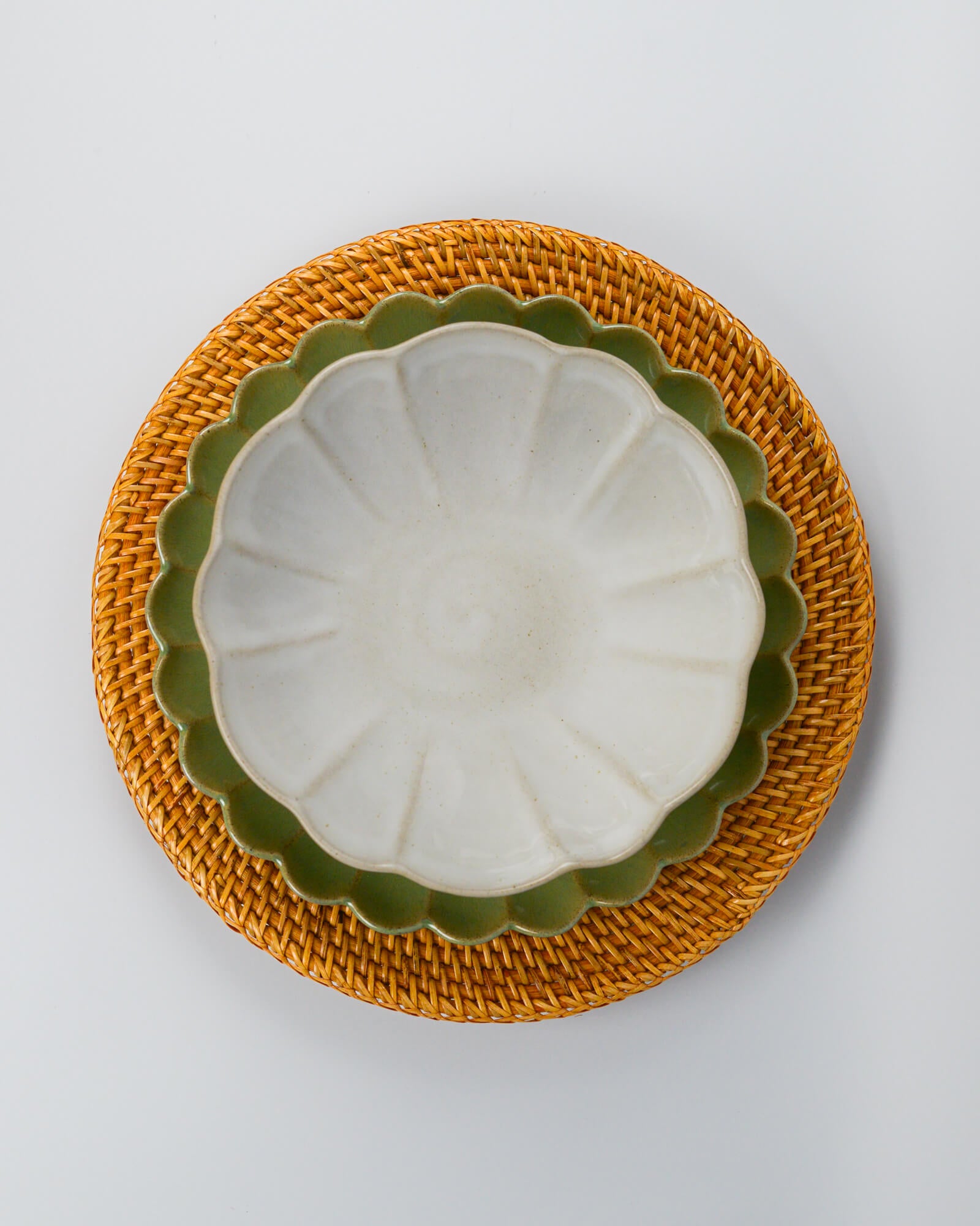 White ceramic pasta bowl layered on a green scalloped edge plate on a rattan charger plate against a white background