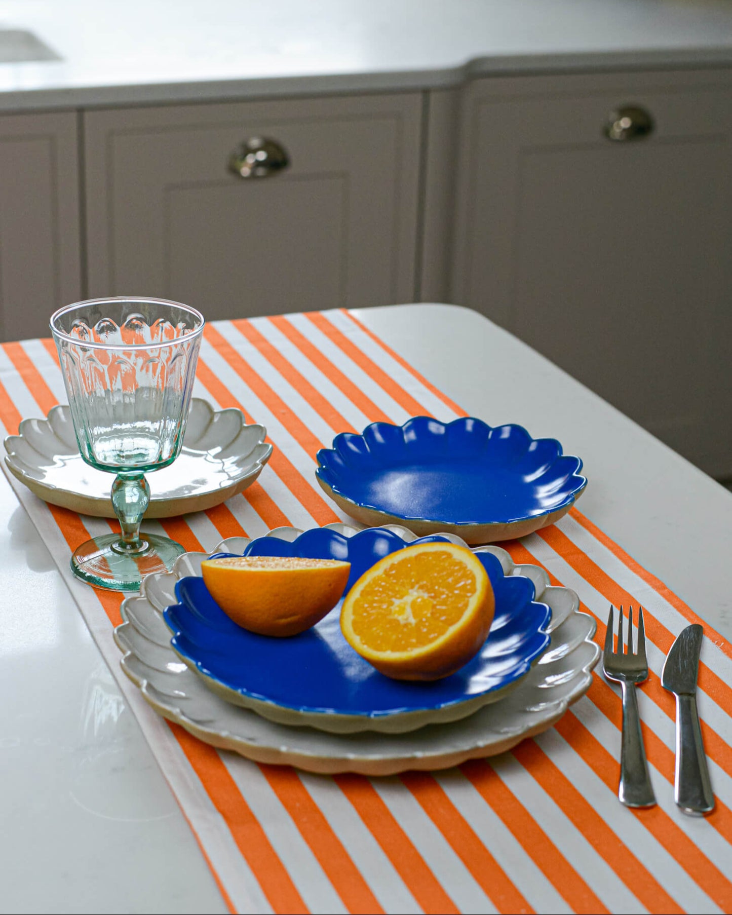 Table setting with blue scalloped edge plates, oranges, a glass, and cutlery on an orange and white striped tablecloth.