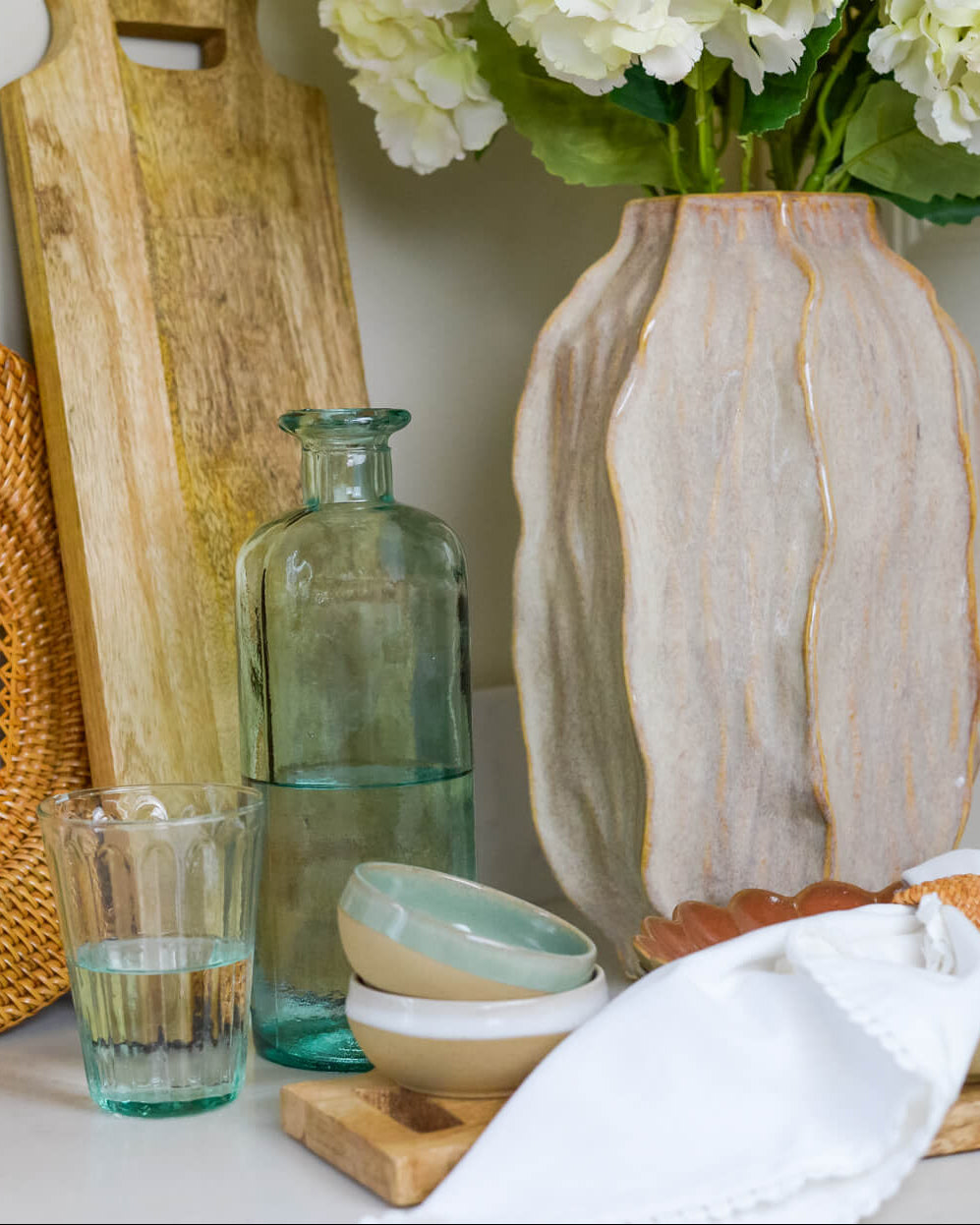 Decorative items including a woven plate, wooden cutting board, glass bottle, and ceramic vase on a surface.