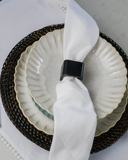 White ceramic scalloped pasta dish with a dark wood napkin ring on a white cotton table runner