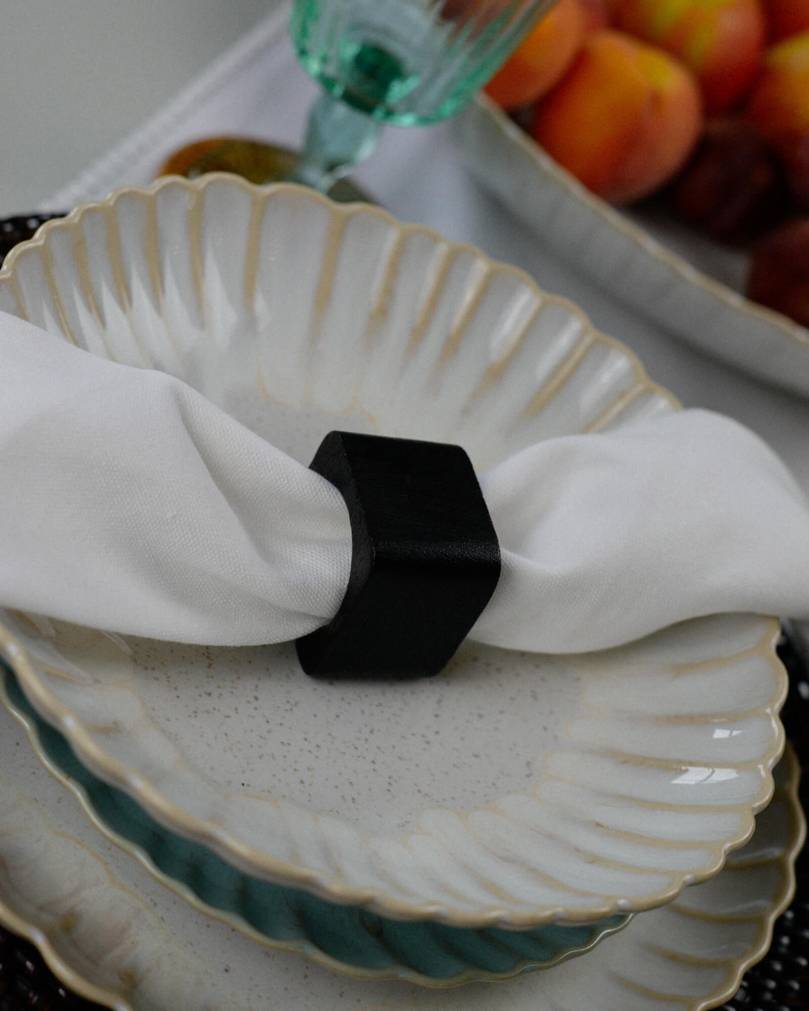 Decorative plate with a white napkin and dark wood napkin ring, with fruits in the background.