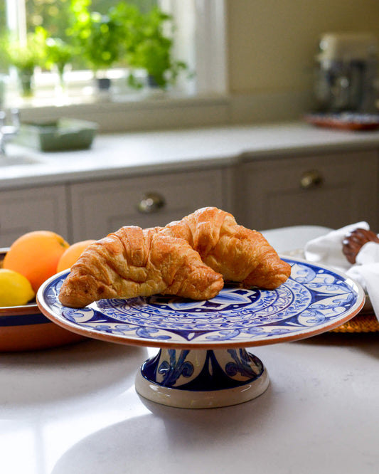Two croissants on a decorative terracotta blue and white footed plate in a kitchen setting.