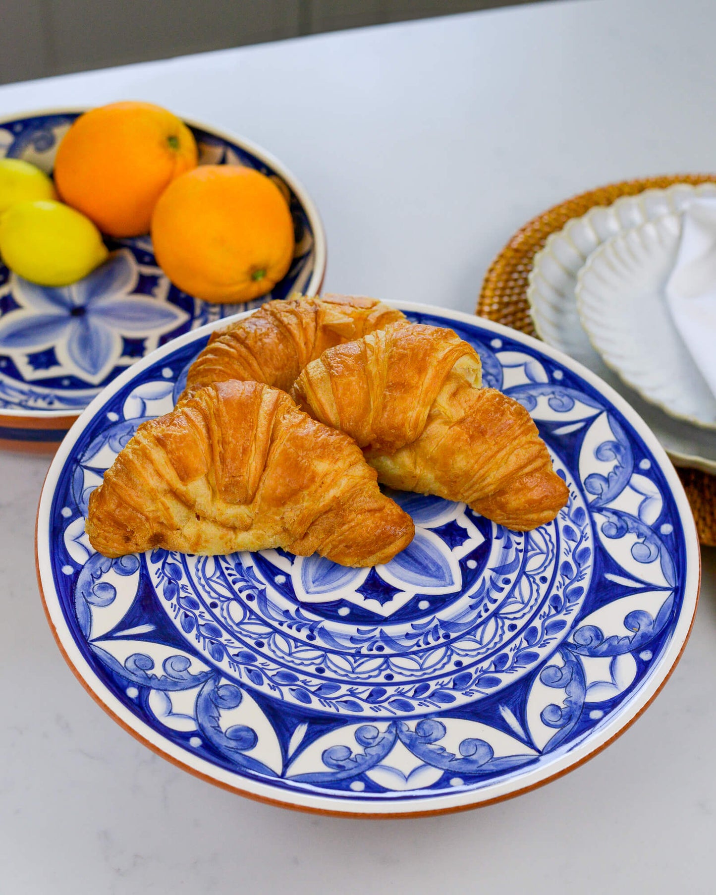 Two croissants on a blue and white decorative footed plate with lemons and oranges in the background.
