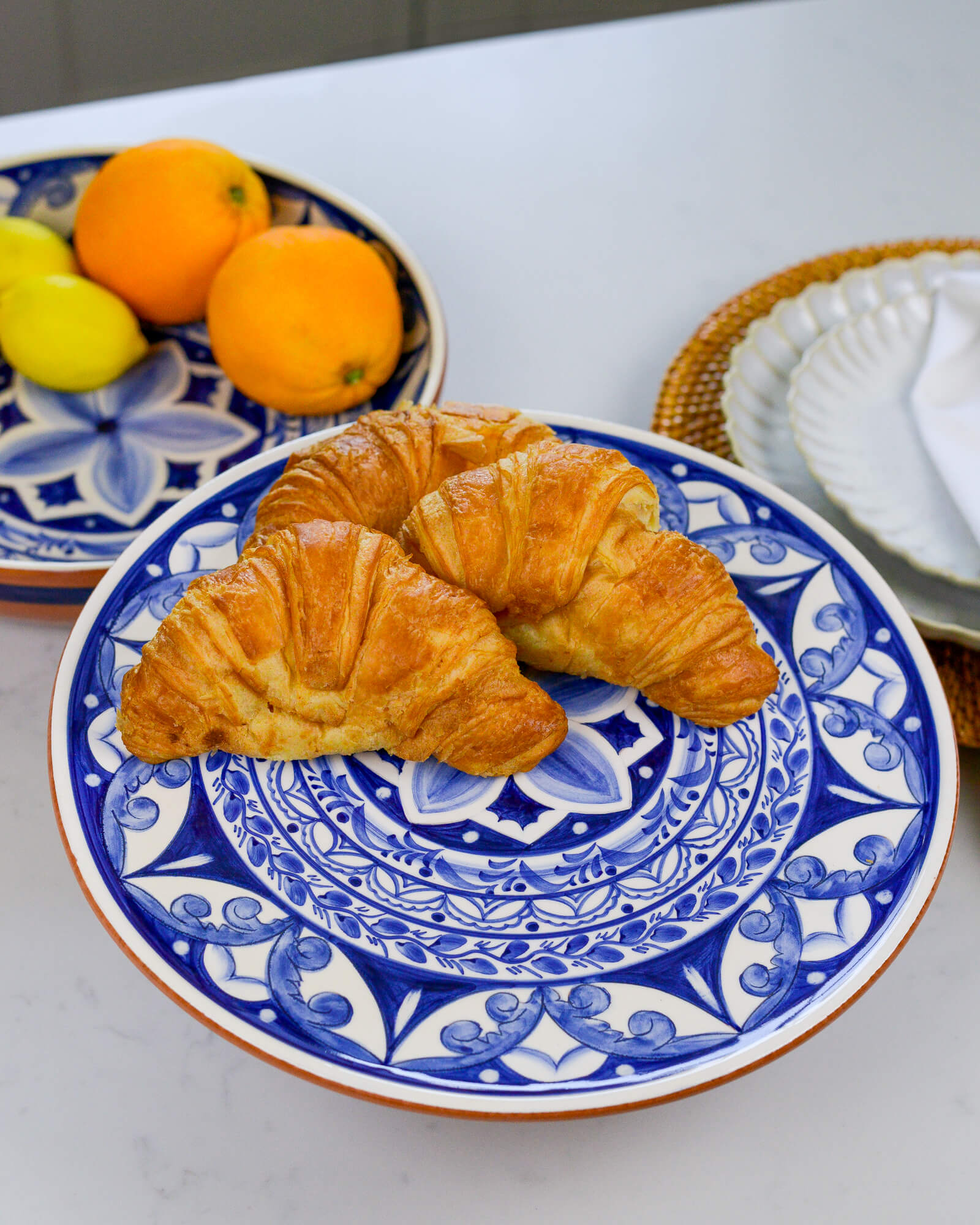 Two croissants on a blue and white decorative footed plate with lemons and oranges in the background.