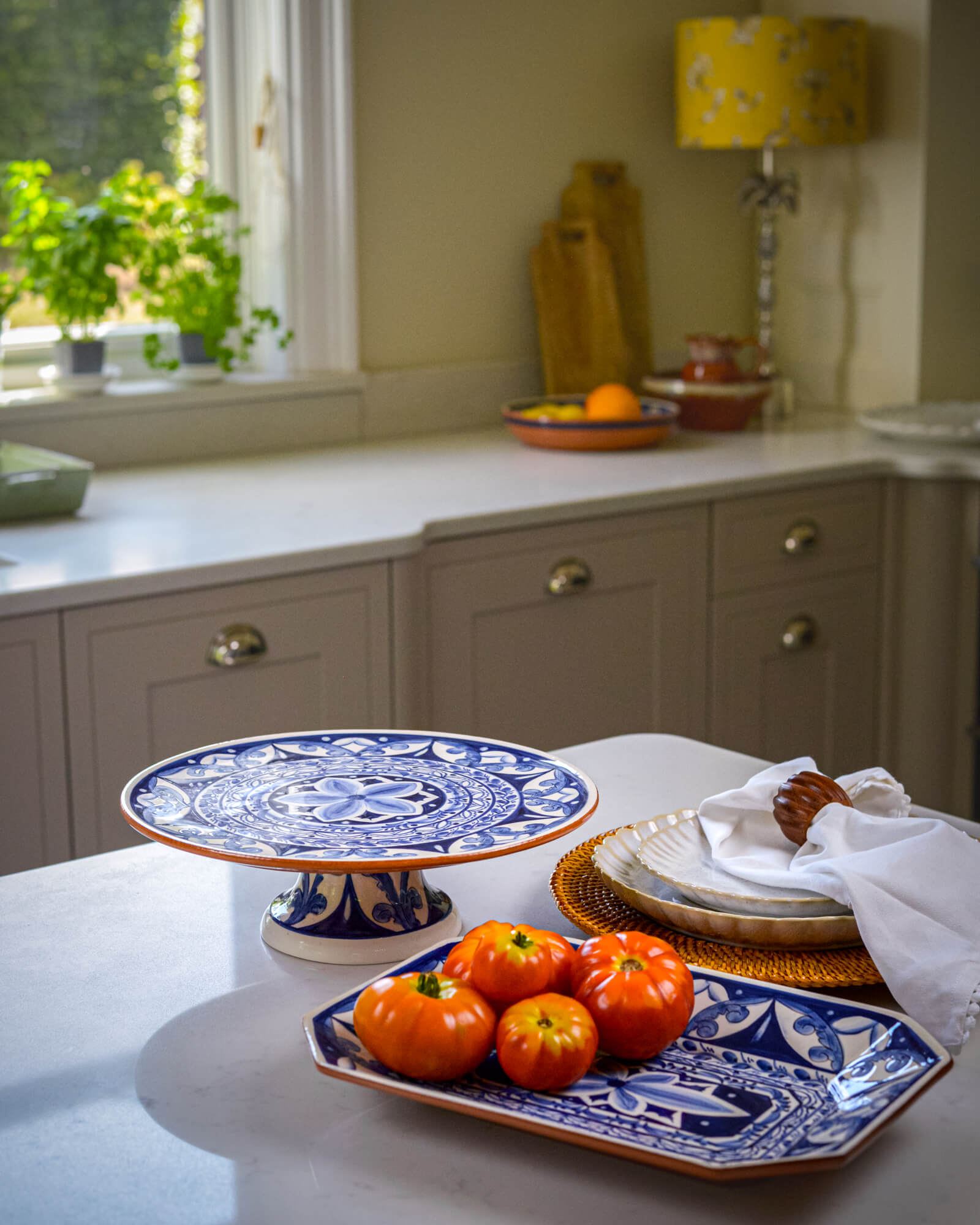 Kitchen counter with blue and white decorative plate and cake stand, tomatoes, and a window in the background.