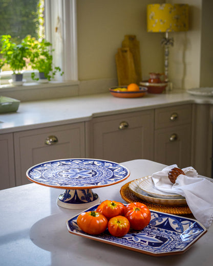 Kitchen counter with blue and white decorative plate and cake stand, tomatoes, and a window in the background.