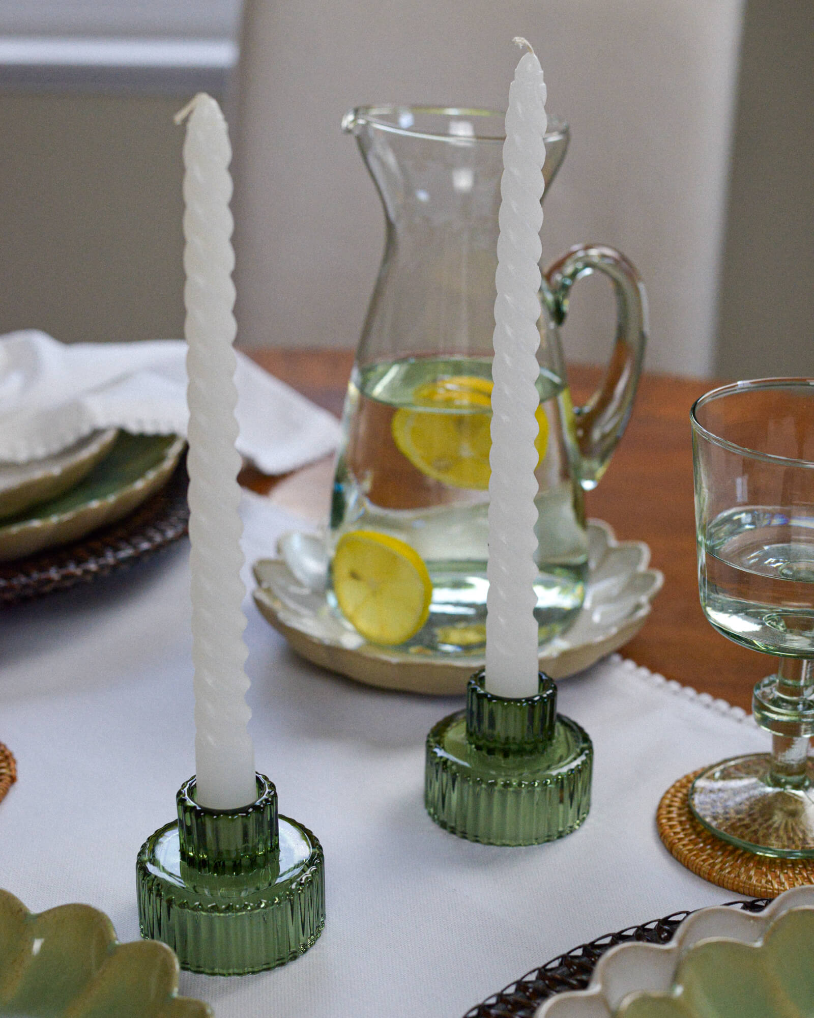 Two green candle holders with white candles on a table setting with a pitcher and glasses.