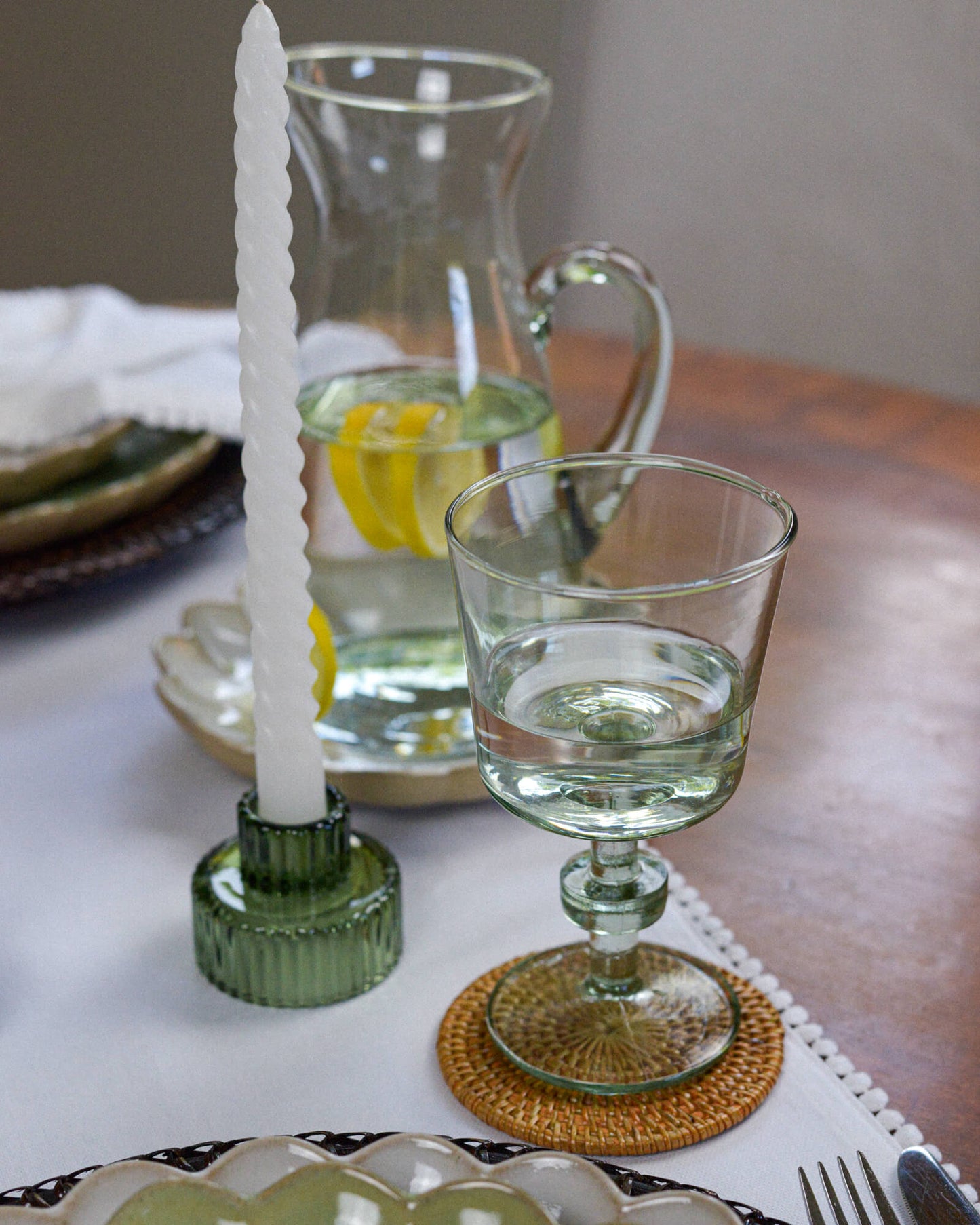 Glass candle holder with a white candle on a table setting with a sculptural wine glass and a recycled glass pitcher.