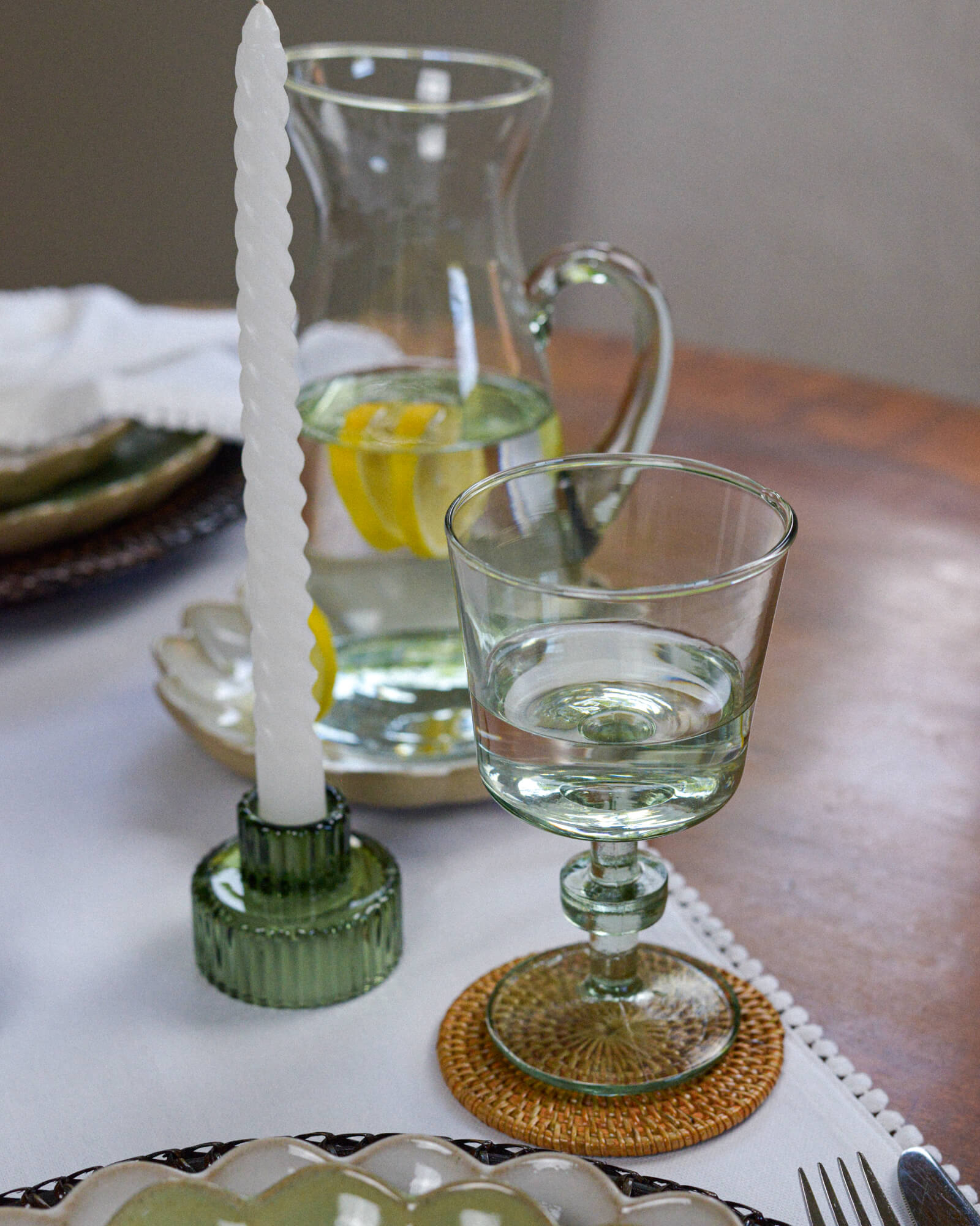 Glass candle holder with a white candle on a table setting with a sculptural wine glass and a recycled glass pitcher.