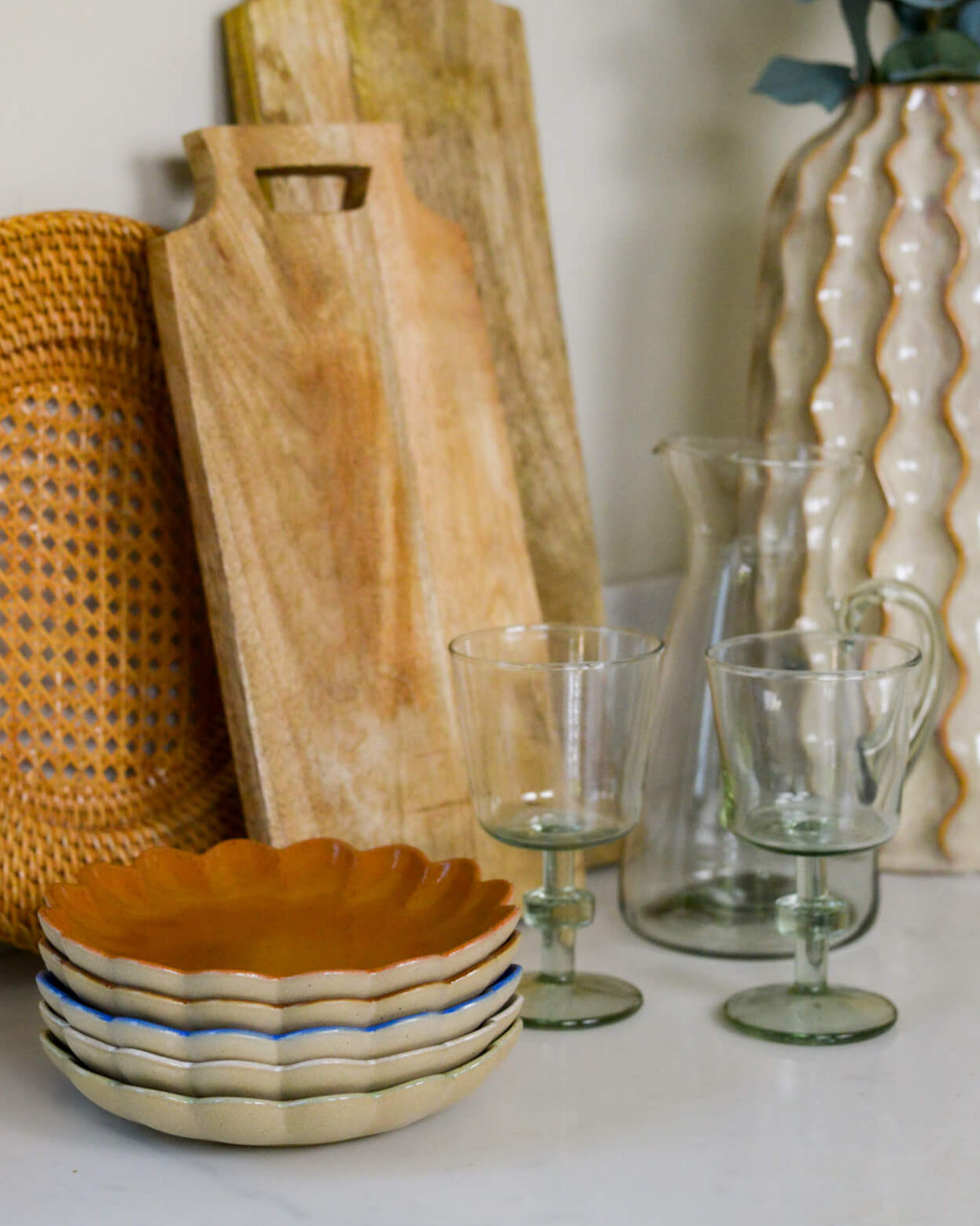 Wooden cutting board, woven plate, ceramic bowl, and glassware on a white marble surface.