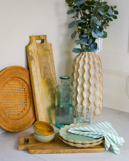 Decorative items including a wooden cutting board, rattan charger plate, ceramic vase, and glass on a light marble surface.