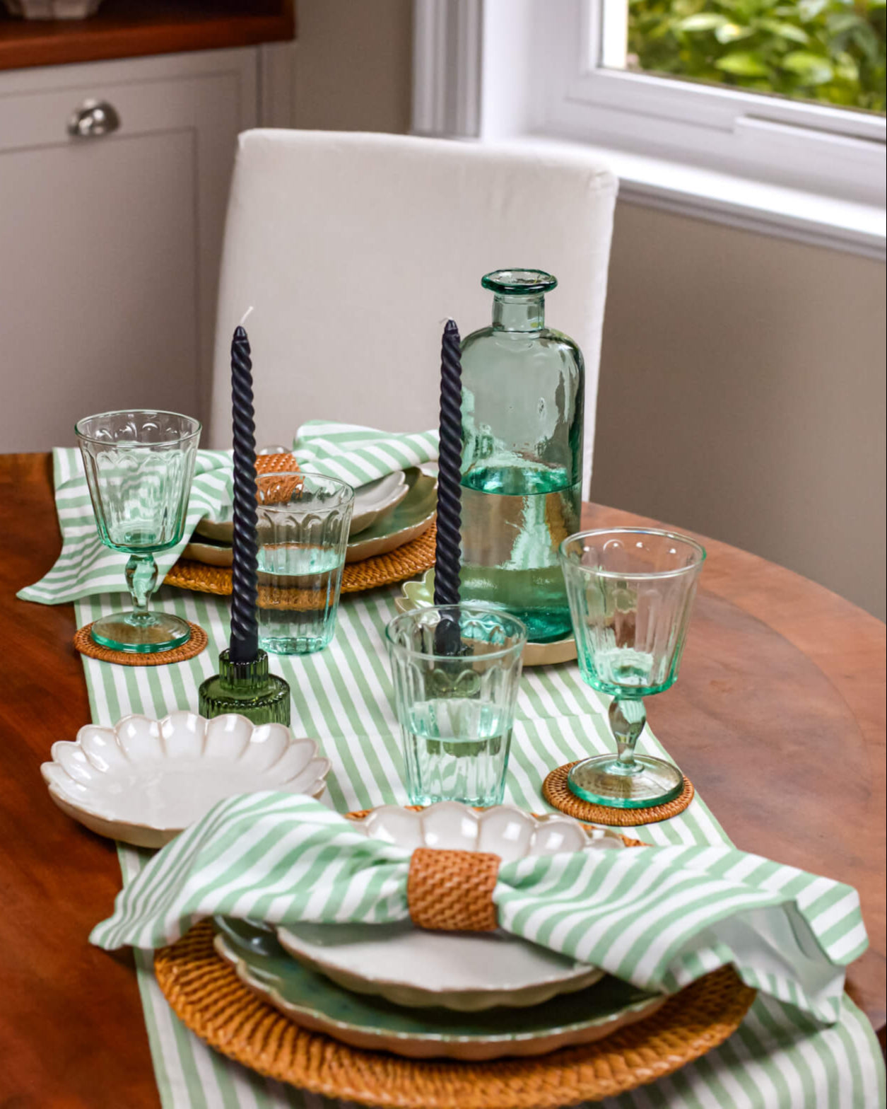 Dining table set with green glasses, candles, and striped napkins and striped green table runner in a home setting.